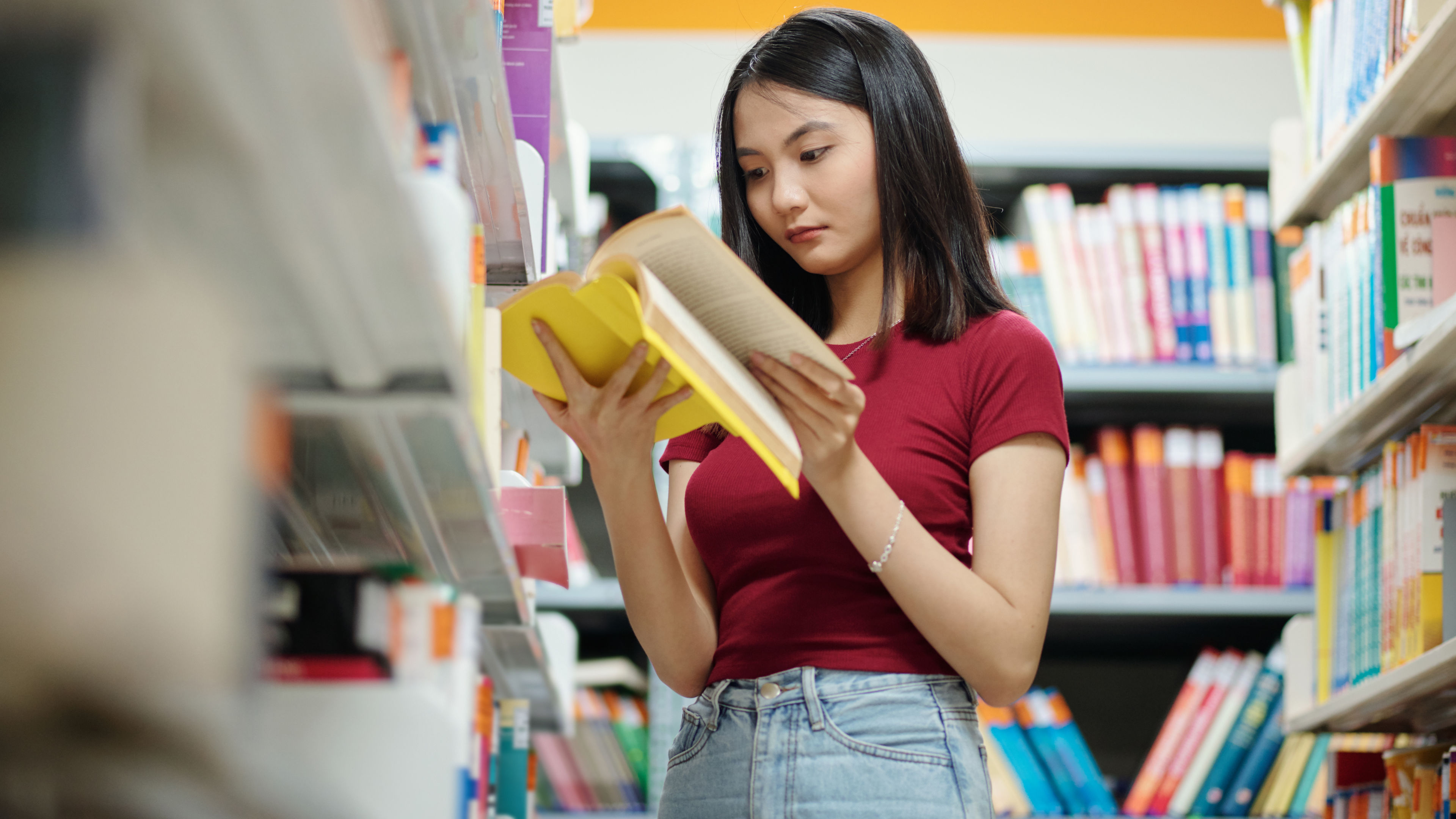 A young woman in a red top and jeans reads a yellow book in a bookstore aisle.