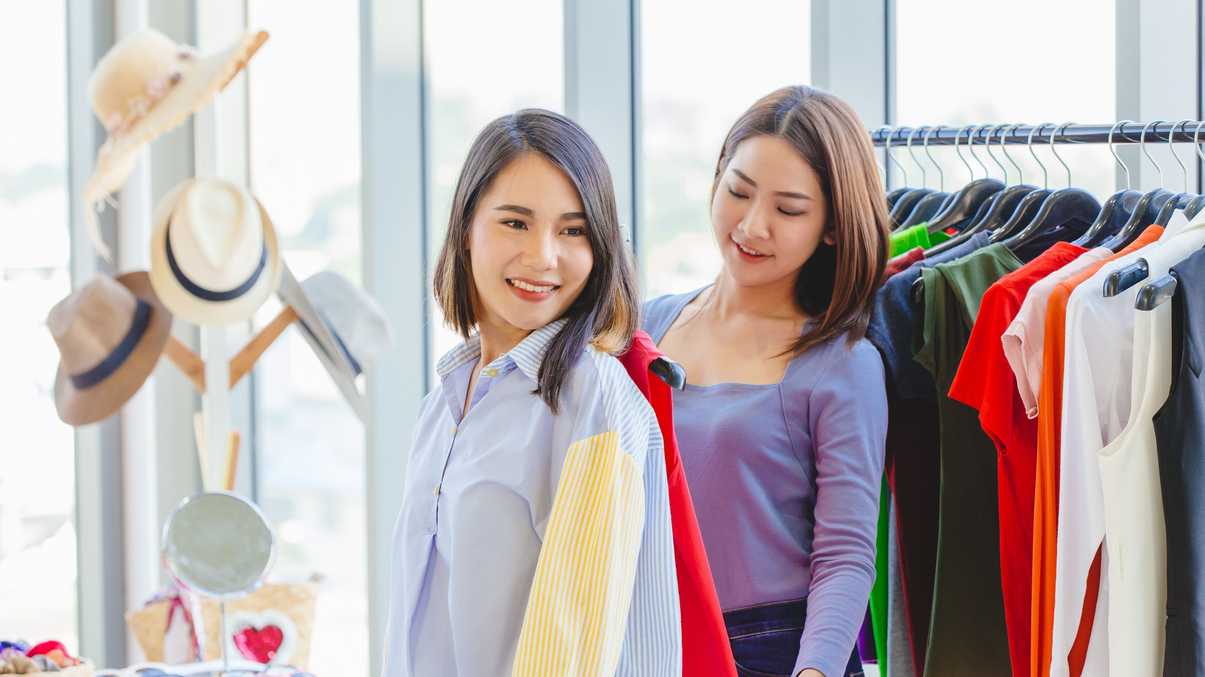 happy women shopping with friend in fashion shop for select new colorful clothes and measure size at shoulder